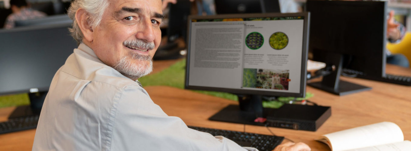 Senior man researching online using computer at a library and looking at the camera smiling - education concepts. **DESIGN ON SCREEN WAS MADE FROM SCRATCH BY US**