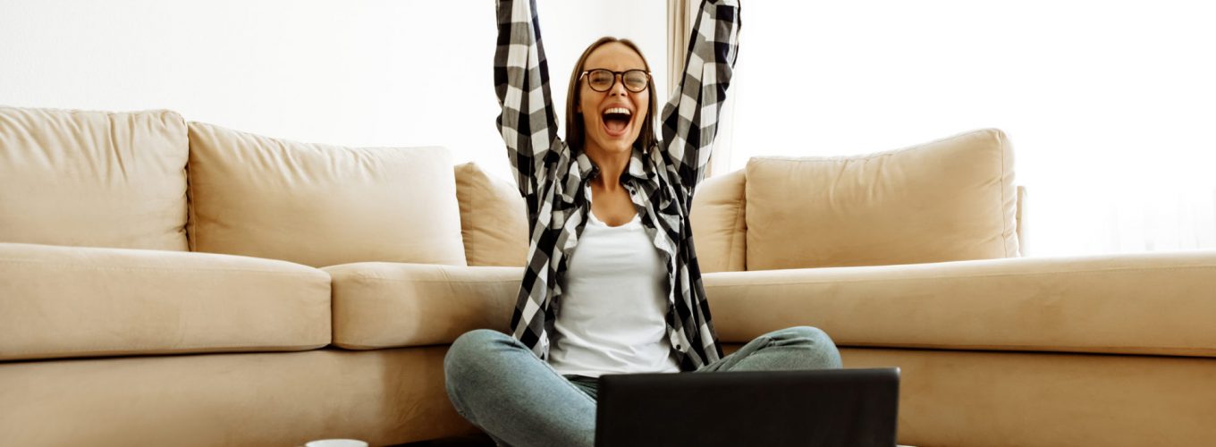 Success. Education. Technology. Home. Girl in casual clothes and eyeglasses is screaming with happiness and raising her hands up while studying with a laptop at home