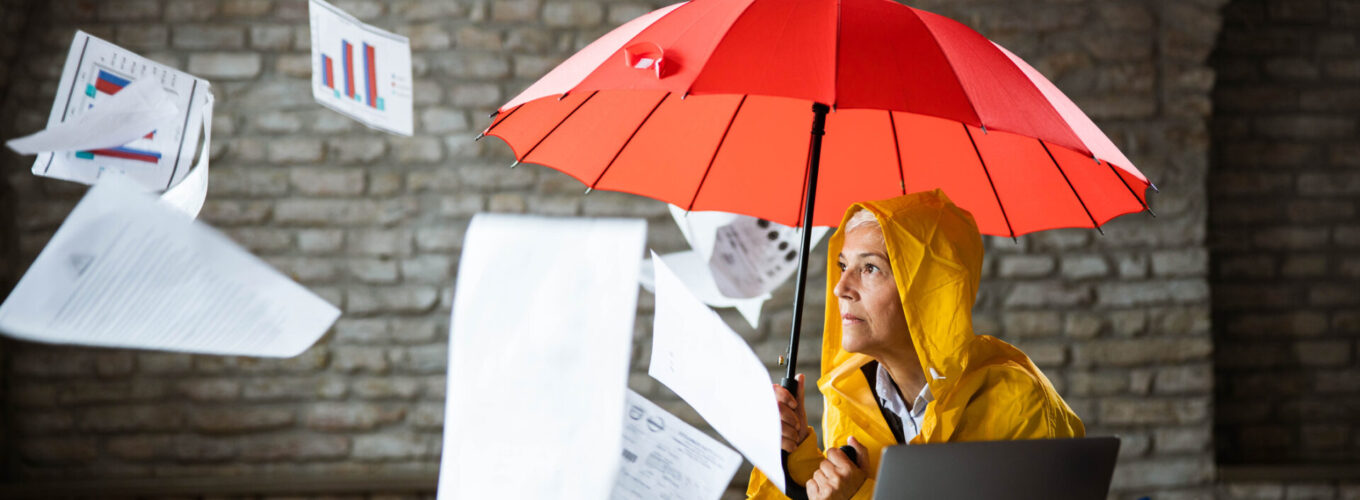 Fearful woman in raincoat holding an umbrella while reports and forms to fill out are falling on her.