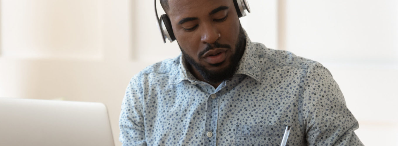 Focused African American student sit at desk wearing wireless headphones learning preparing for seminar or exam, guy interpreter hears audio writing down translation, online lecture course e-learning concept