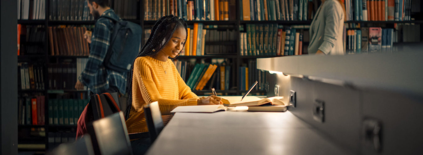 University Library: Gifted Black Girl uses Laptop, Writes Notes for the Paper, Essay, Study for Class Assignment. Students Learning, Studying for Exams College. Side View Portrait with Bookshelves