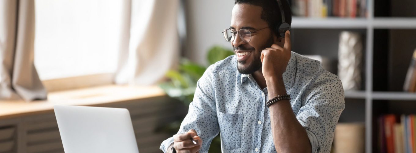 African guy student learn on-line use headphones with mic and pc holds pen takes notes in notebook sit at desk at home. Businessman wear headset talk by conference call do video chat working at office