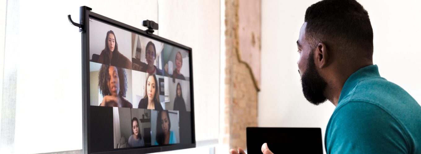 A mid adult man gestures while talking with a group of colleagues during a virtual staff meeting via video conference.