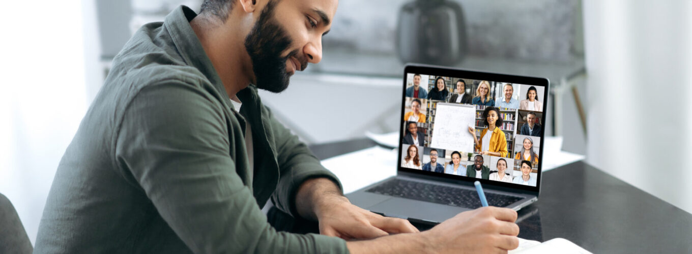 Side view of a successful smart guy listening to an online lecture, taking notes in a notebook, on a laptop screen, a teacher and a group of multiracial people. Online training, webinar