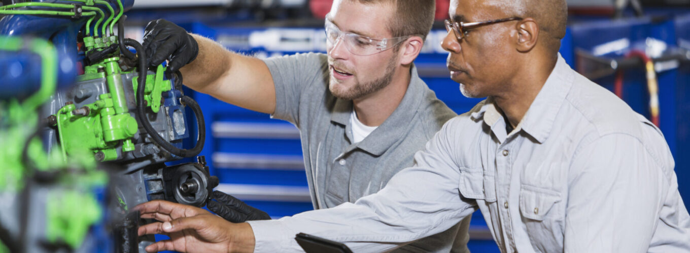 An African American man instructing a Caucasian student on how to repair a diesel engine. He is teaching a class in a vocational school, for learning the auto mechanic trade. They are kneeling by an engine block which is color-coded for the classroom.