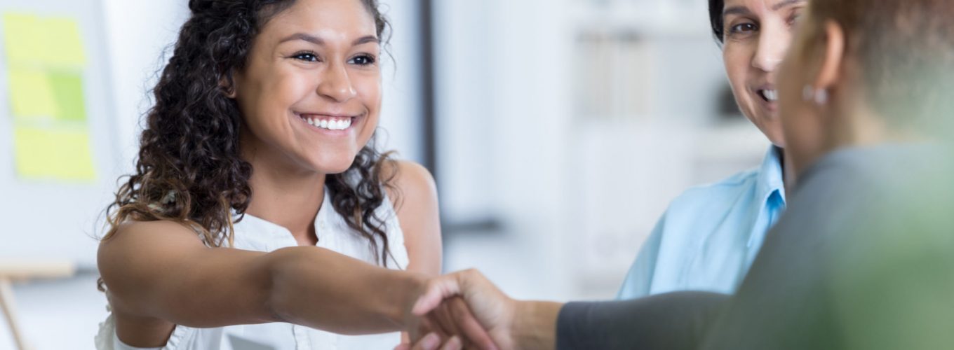 Confident young woman interviews for a position in an office. She is greeting the office manage. She is smiling confidently.