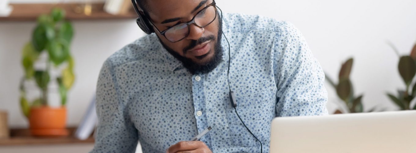 Focused african business man in headphones writing notes in notebook watching webinar video course, serious black male student looking at laptop listening lecture study online on computer e learning