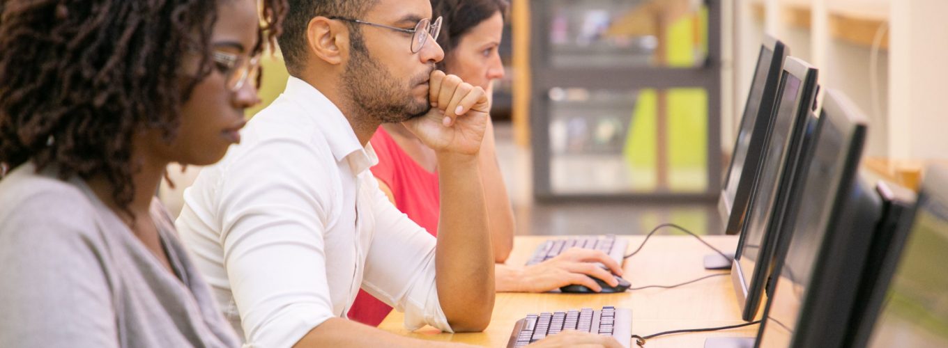 Multiracial group of students training in computer class. Line of man and women in casual sitting at table, using desktops, typing, looking at monitor. Training center concept