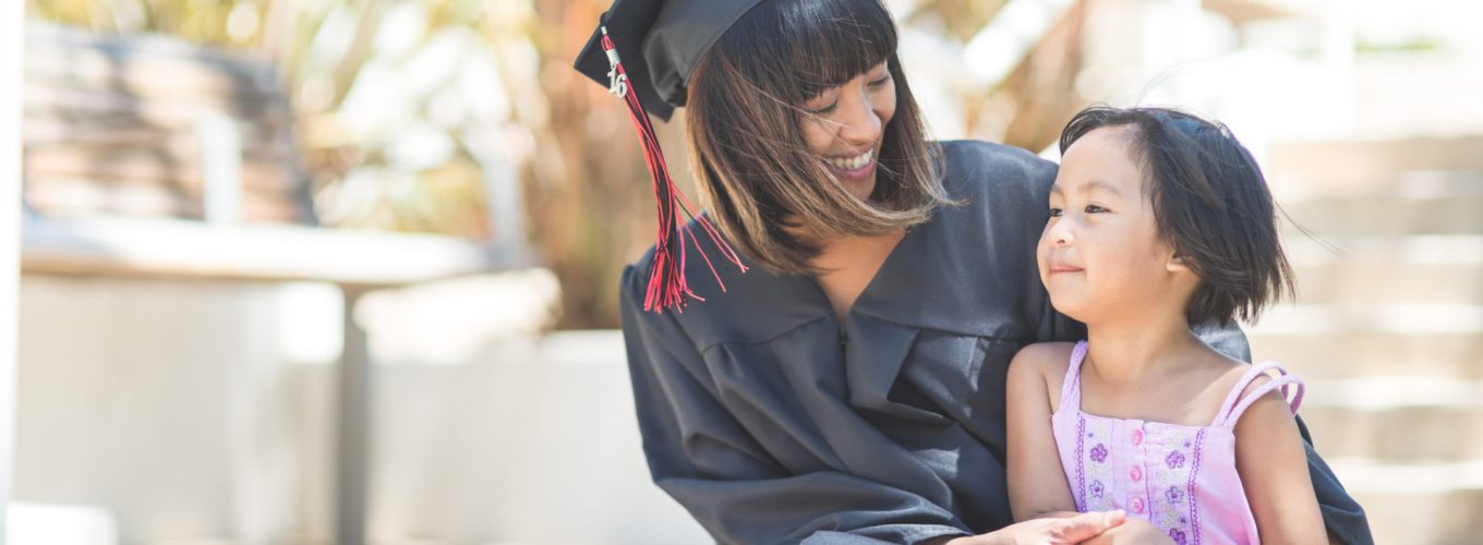 Attractive Filipino woman in graduation robe and cap sits on steps with her young daughter after graduation. They are looking at each other affectionately and holding hands.